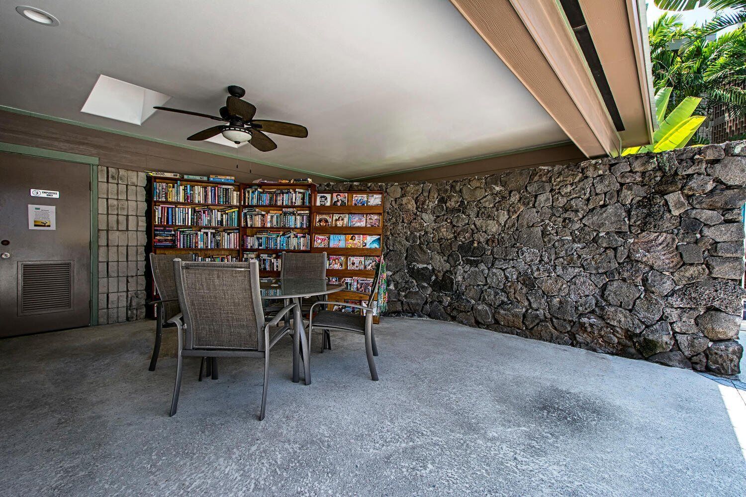 Una habitación con pared de piedra, una estantería llena de libros, un ventilador de techo y una puerta con un letrero.