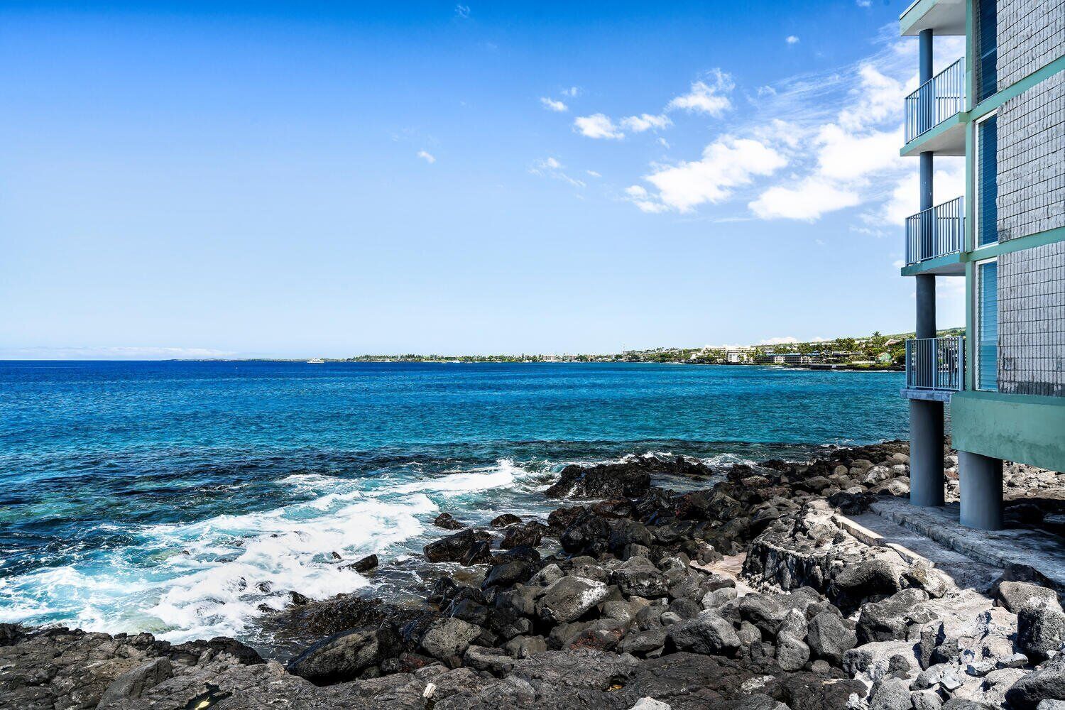 A coastal view with a rocky shoreline, a building with balconies overlooking the sea, and a distant shoreline with buildings.