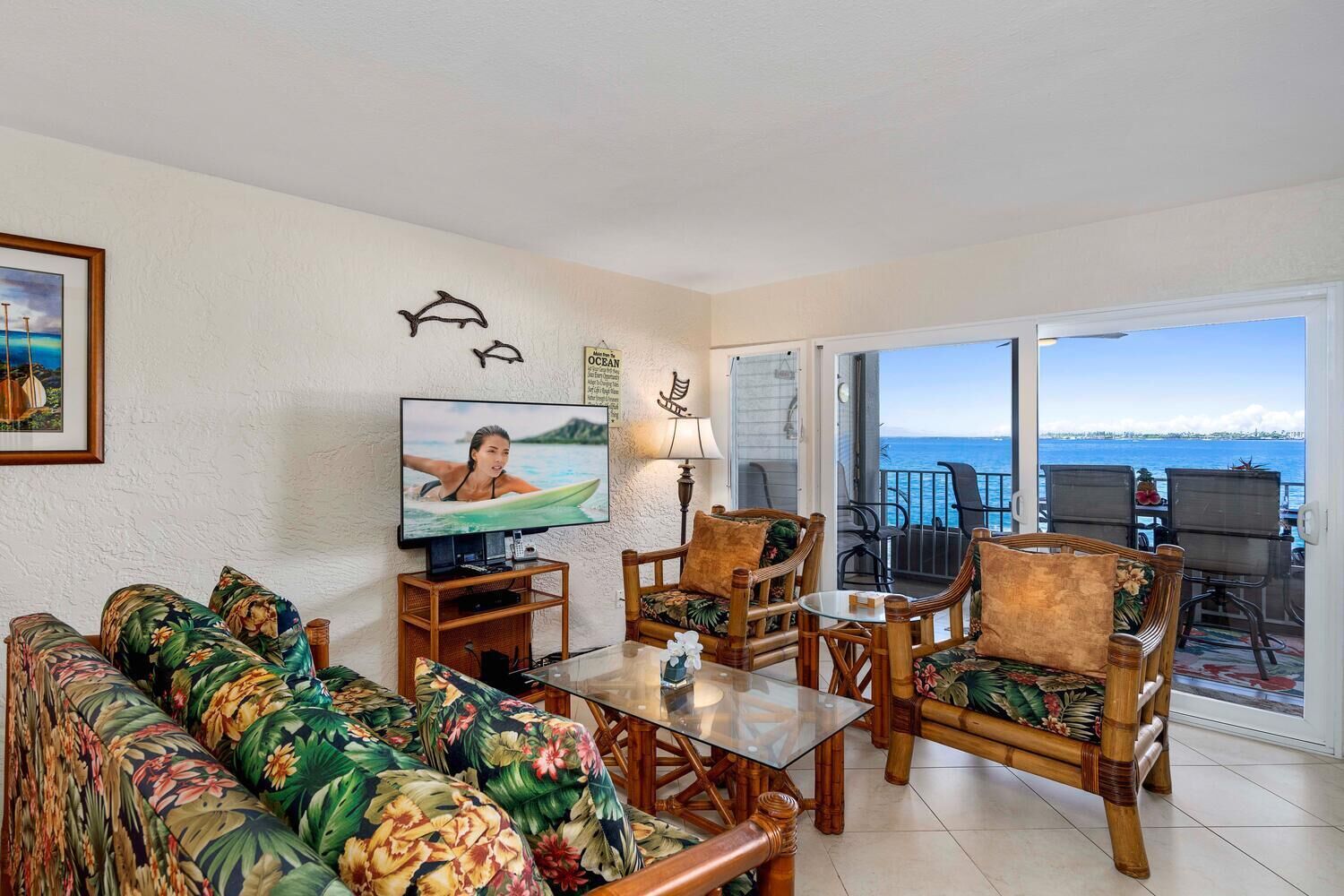 Ein Wohnzimmer mit Fernseher, Sofa, Sesseln und einem Glastisch. Es gibt einen Balkon mit Meerblick.