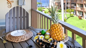Une table en bois sur laquelle se trouvent une assiette, un ananas, un citron vert et des fleurs, le tout placé sur un balcon donnant sur une piscine et des bâtiments.