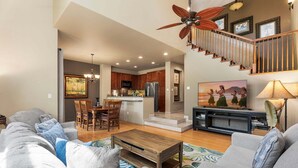 A spacious living area with a sofa, coffee table, and a TV. There's a kitchen in the background with a dining area and a staircase. - Beautifully Furnished - Hawaiian Villa - AC & Wifi! - Vrkcv (Waikoloa)