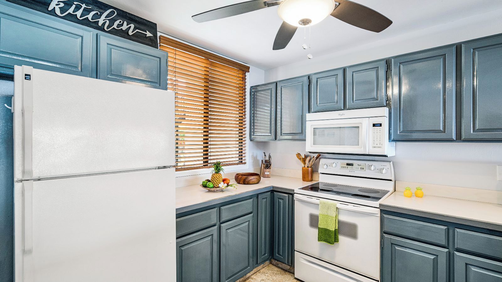 A kitchen with blue cabinets, a white refrigerator, a microwave, an oven, and a ceiling fan.
