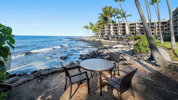 A beachfront patio with a round table and chairs, overlooking the ocean with palm trees and a hotel building in the background.