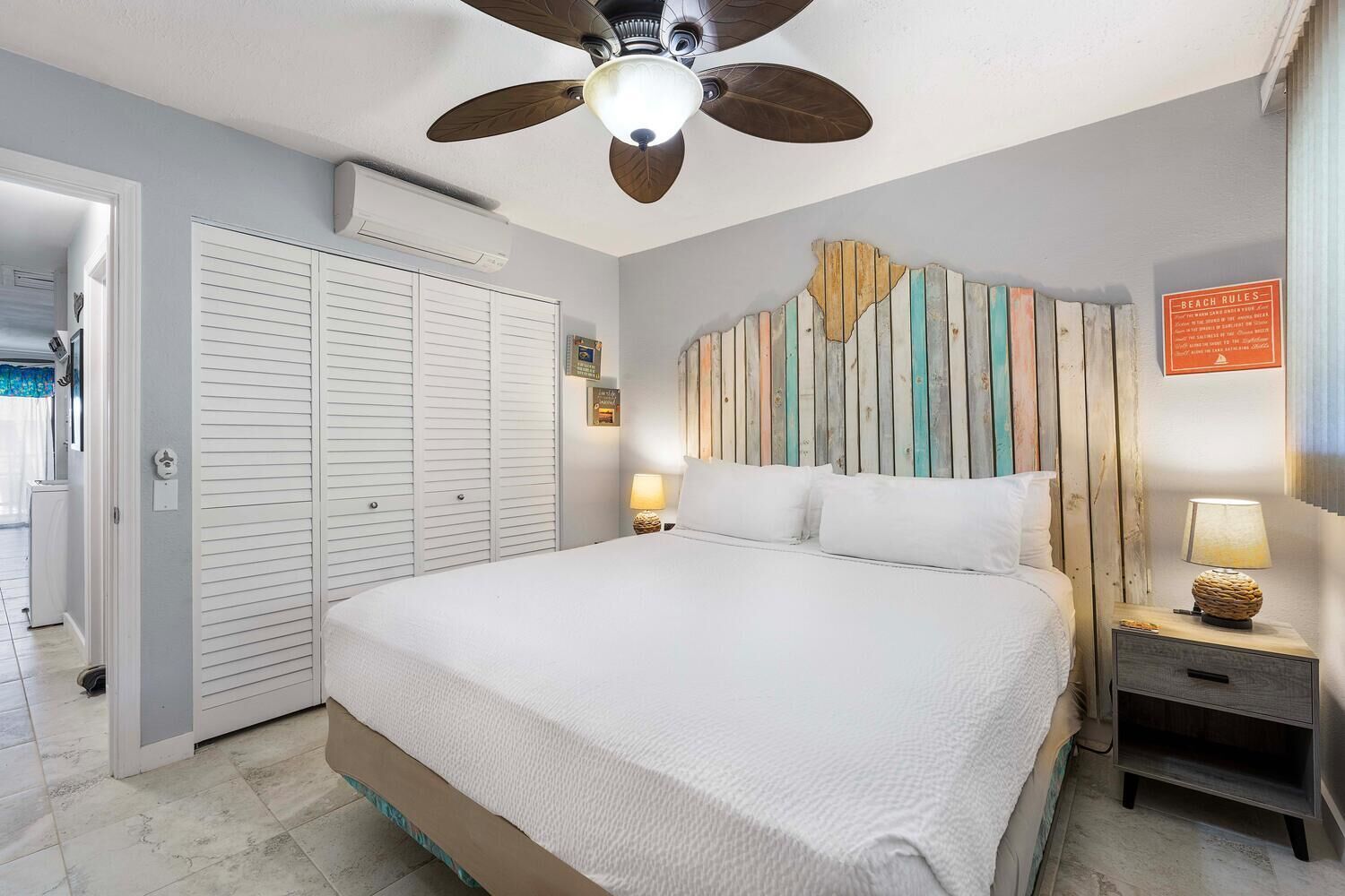 Un dormitorio con cama, ventilador de techo, aire acondicionado de pared, cabecero de madera, lámparas de noche y un letrero sobre normas de la playa.