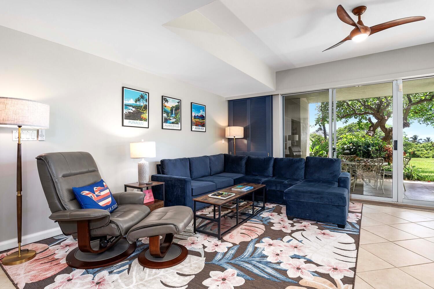 Un salon comprenant un canapé, un fauteuil, une table basse et un ventilateur de plafond. Des tableaux encadrés ornent le mur, et une porte vitrée offre une vue sur l’extérieur.