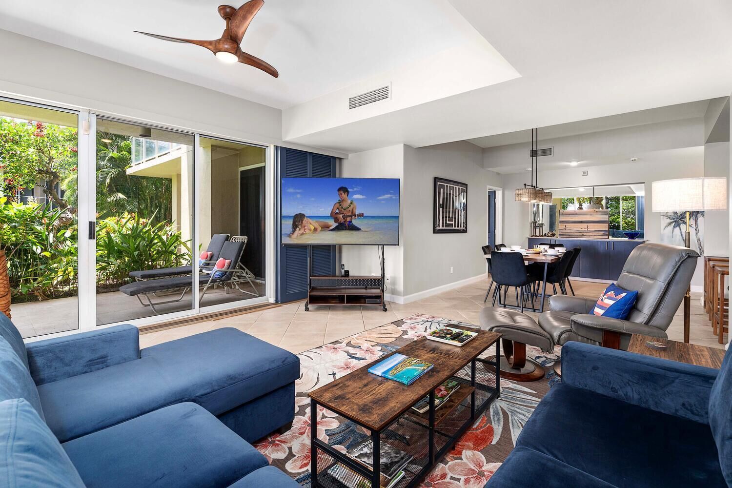 A modern living room with a large TV, blue sofas, a dining area, and a ceiling fan.
