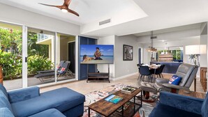 A modern living room with a large TV, blue sofas, a dining area, and a ceiling fan. - Ground Floor Corner Unit - Vrkcv (Waikoloa)