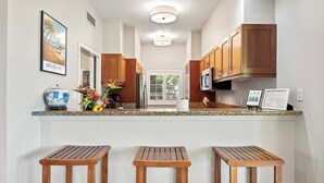 A kitchen with wooden cabinets, a granite countertop, and three wooden stools. - Ground Floor Corner Unit - Vrkcv (Waikoloa)