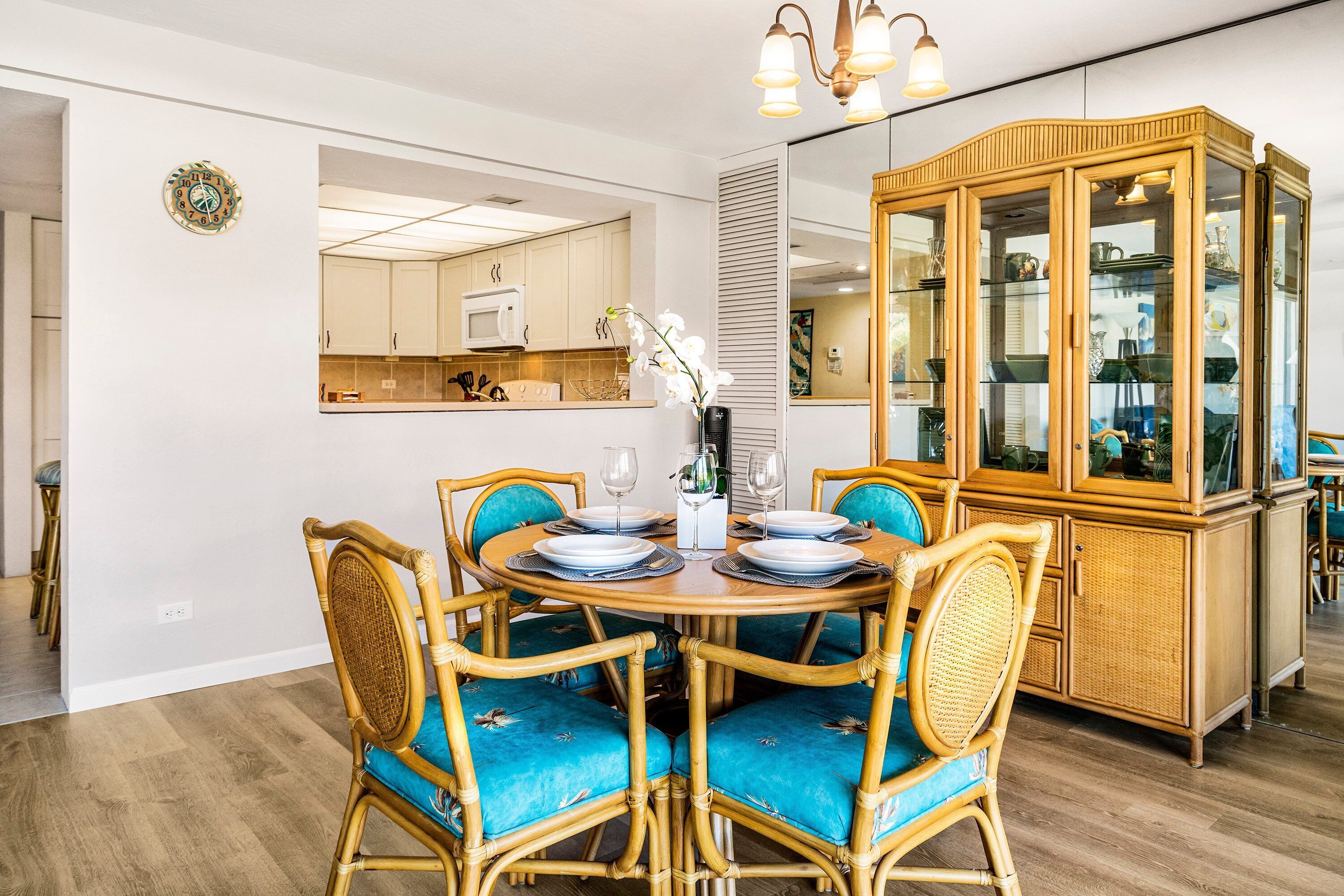 Un comedor con una mesa redonda preparada para cuatro personas, rodeada de sillas de mimbre con cojines azules. A la derecha, un mueble de madera con puertas de cristal.