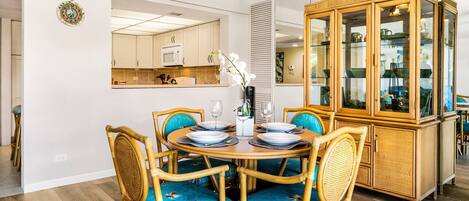Un comedor con una mesa redonda preparada para cuatro personas, rodeada de sillas de mimbre con cojines azules. A la derecha, un mueble de madera con puertas de cristal.