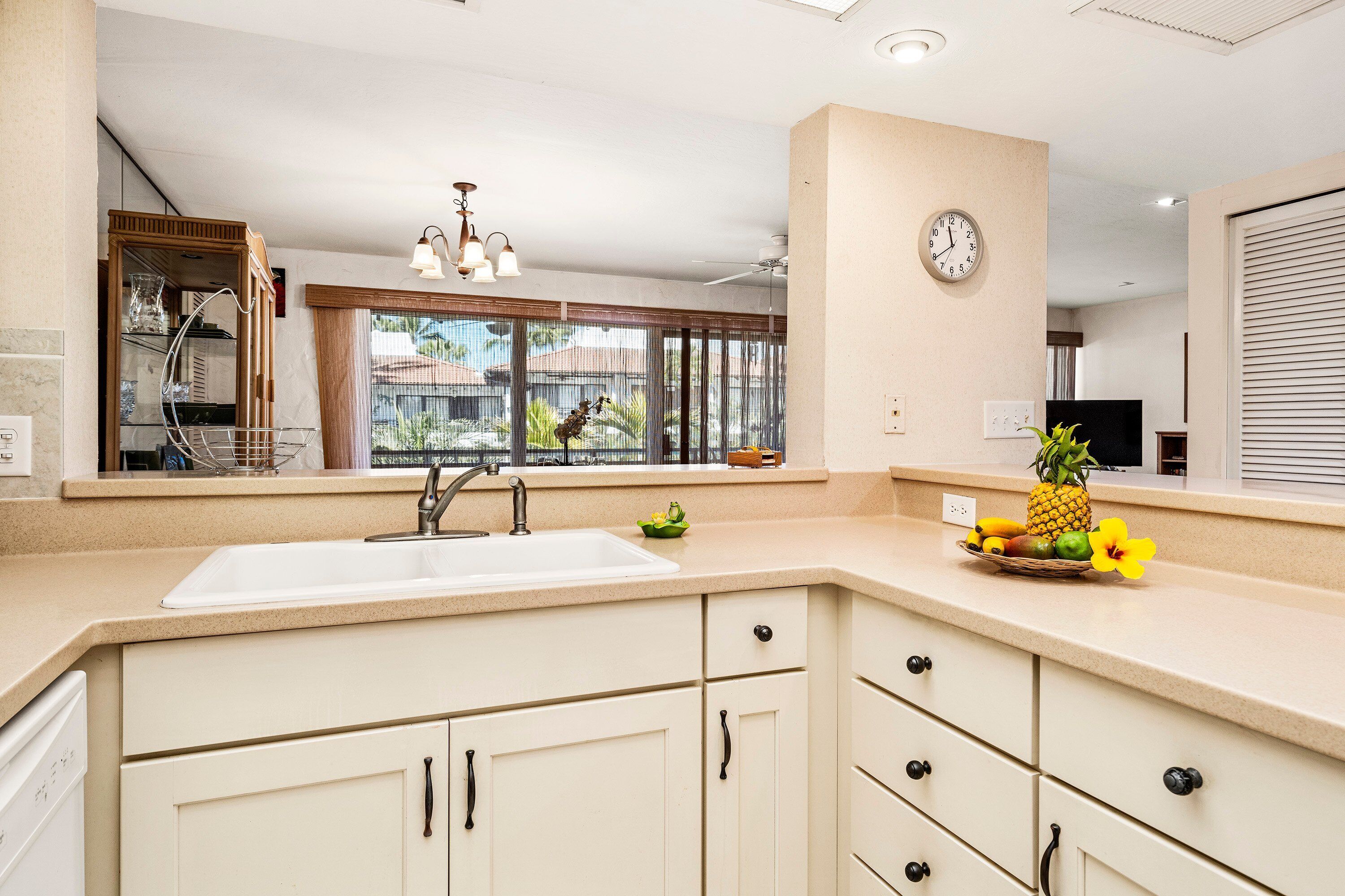 A kitchen with a sink, cabinets, a clock, and a window with blinds.