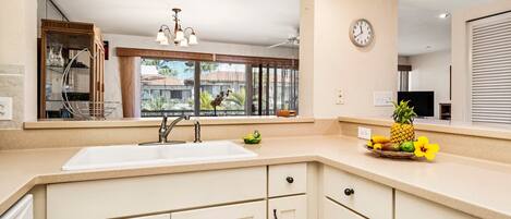 A kitchen with a sink, cabinets, a clock, and a window with blinds.