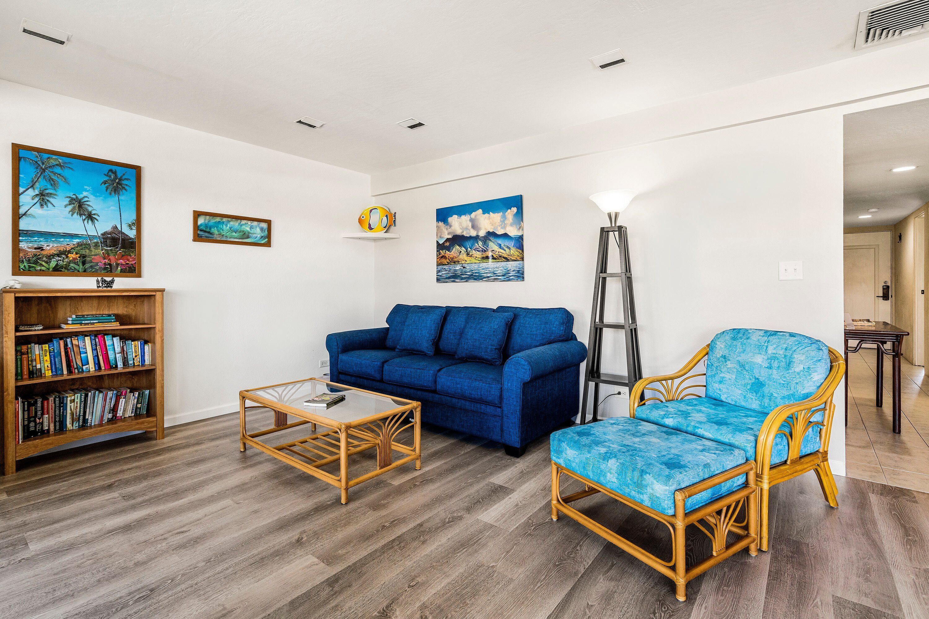 Una sala de estar con un sofá azul, una estantería de madera, un sillón de mimbre y dos cuadros en la pared.