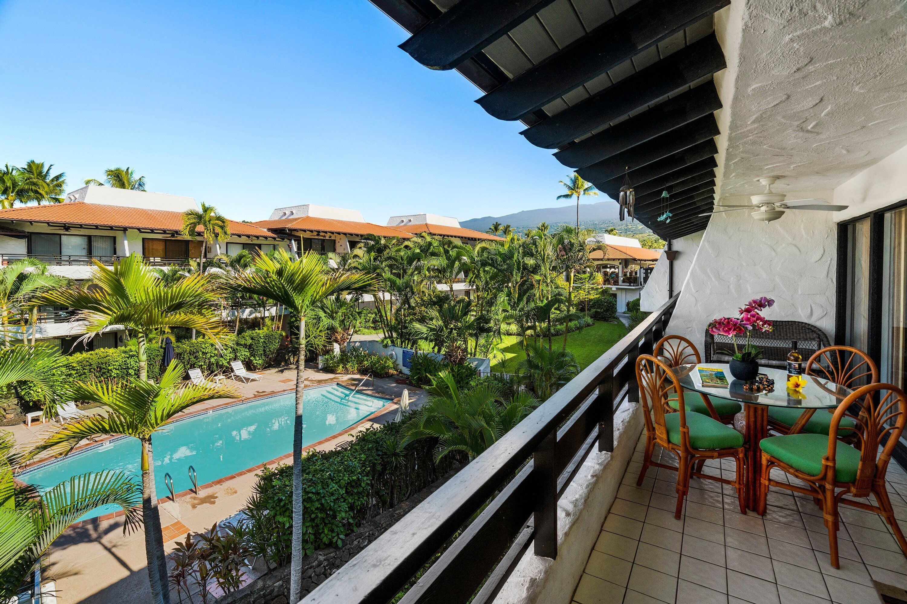 A balcony with a view of a swimming pool, surrounded by palm trees and a building with a tiled roof.