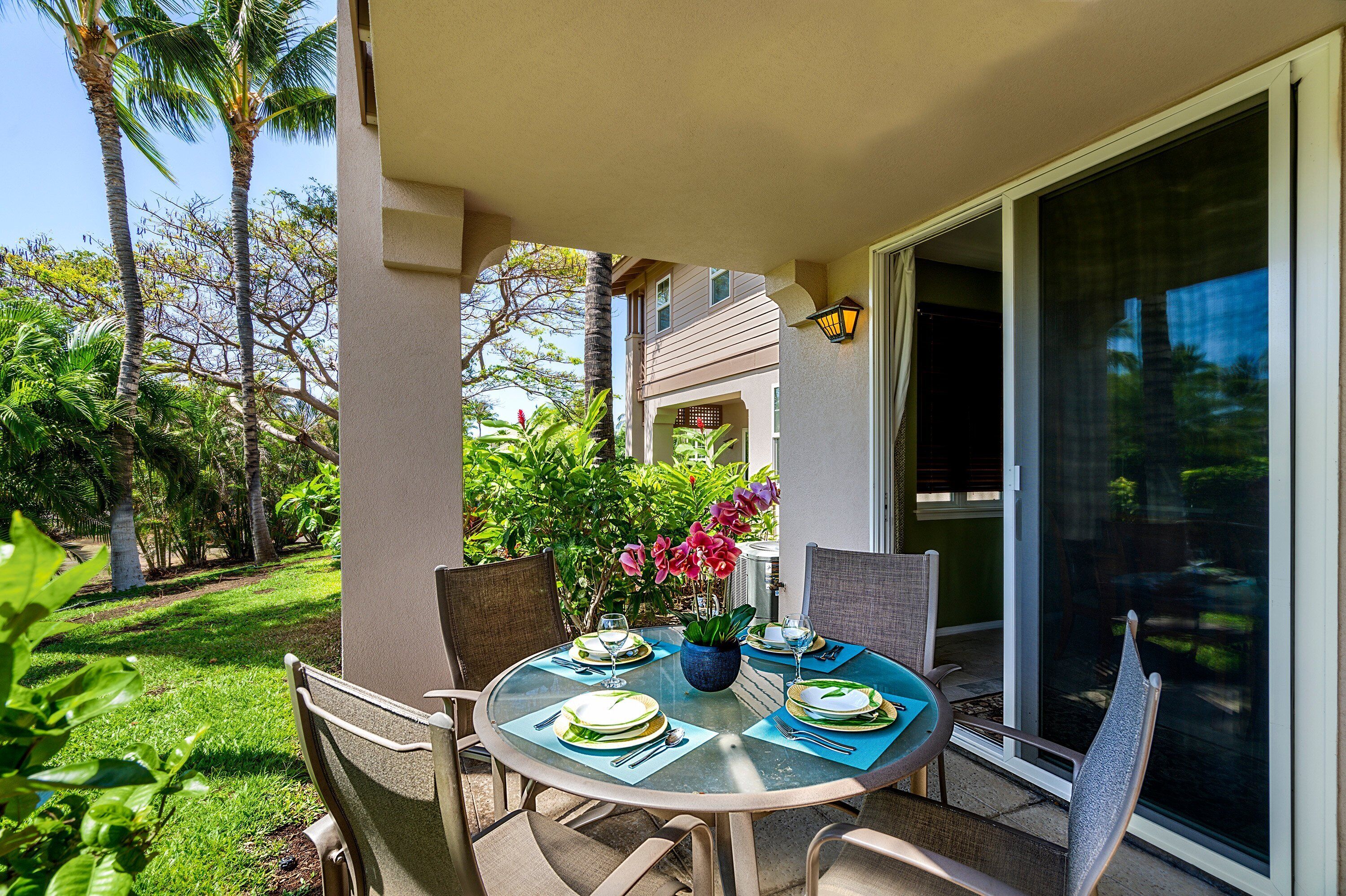 A round glass table set for dining with chairs, a potted plant, and a view of a lush garden and building in the background.