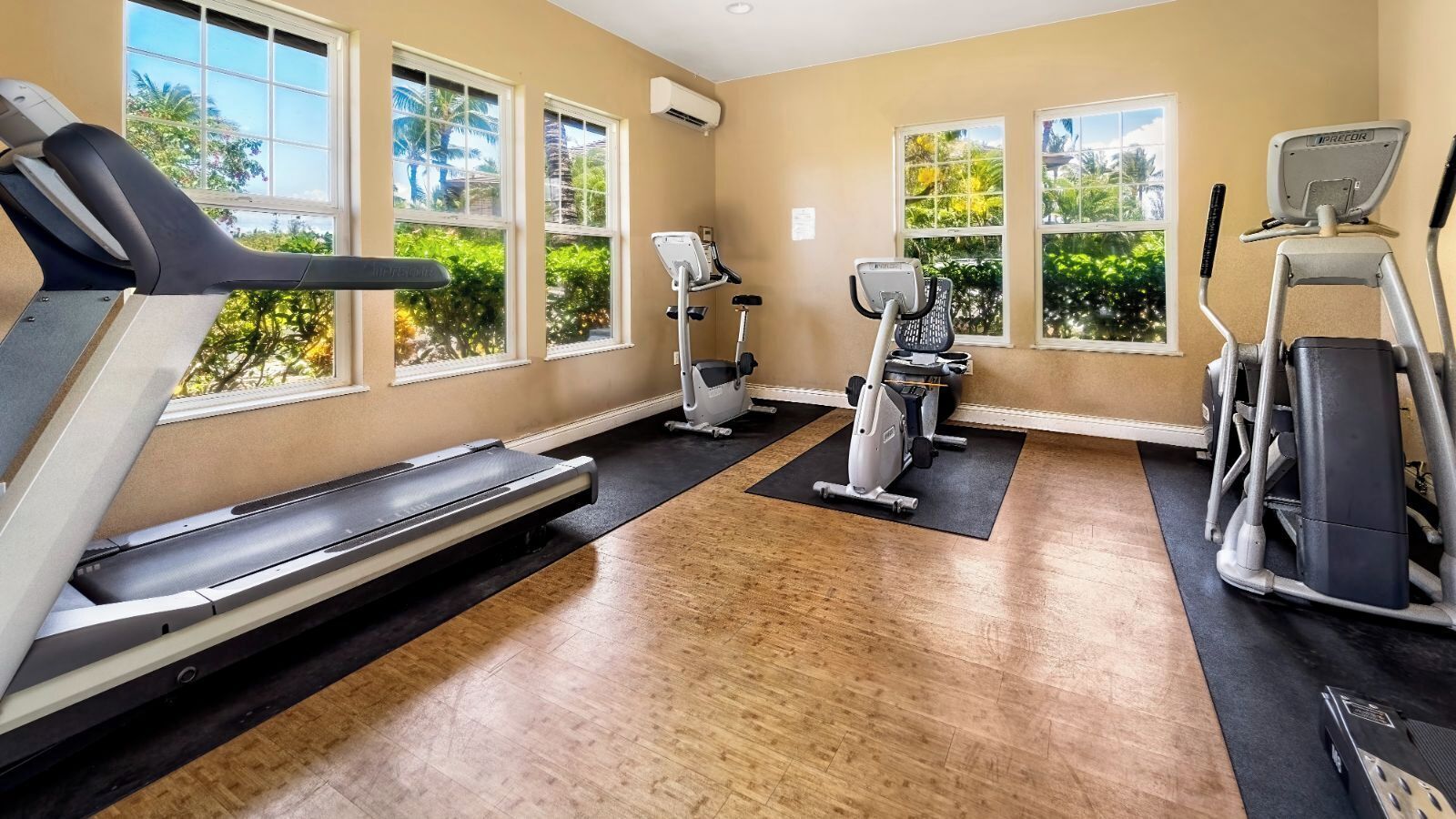 Ein Fitnessstudio mit Laufbändern, Ergometer und Crosstrainern, großen Fenstern mit Blick auf Grünflächen und Holzböden.