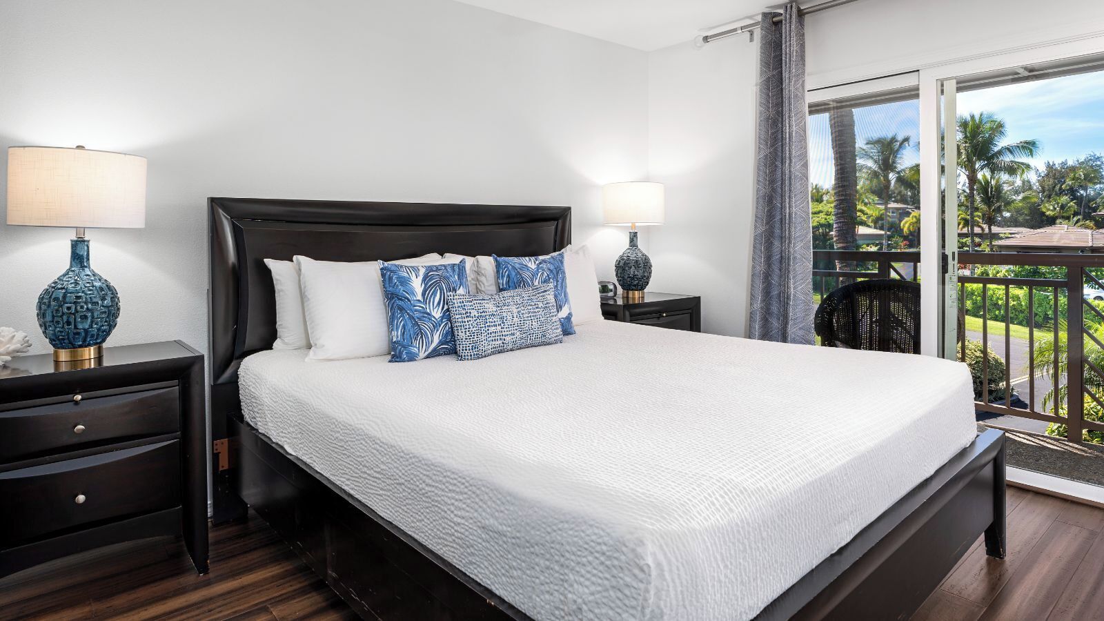 A bedroom with a large bed, two bedside tables with lamps, a dark wooden headboard, and a balcony with a view of palm trees.