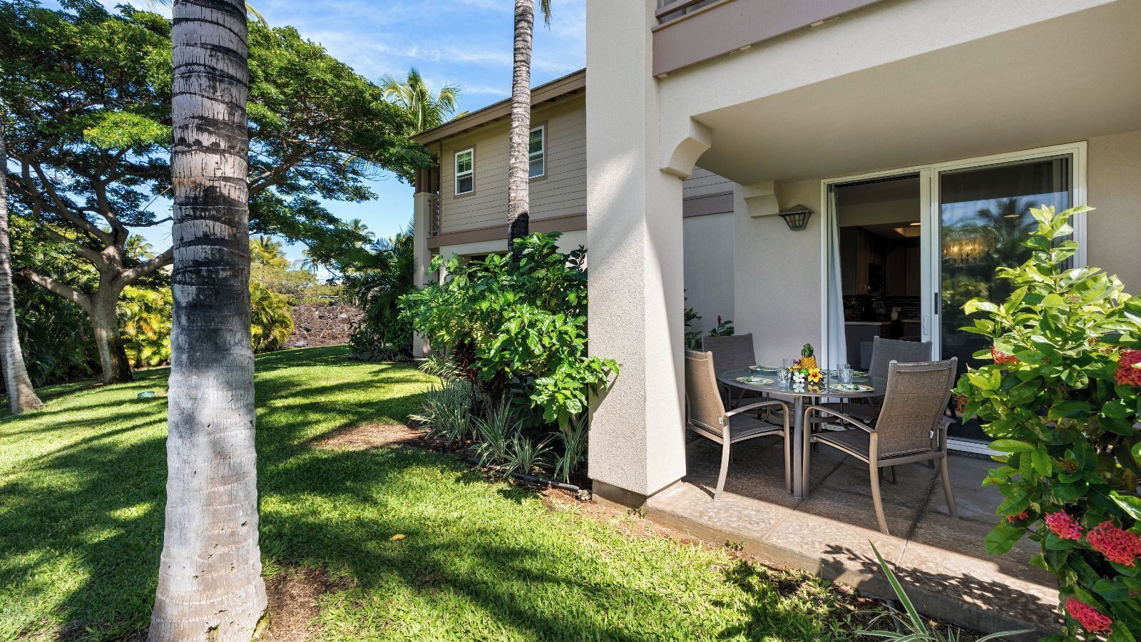 A house with a patio, a dining table with chairs, and a garden with palm trees and shrubs.