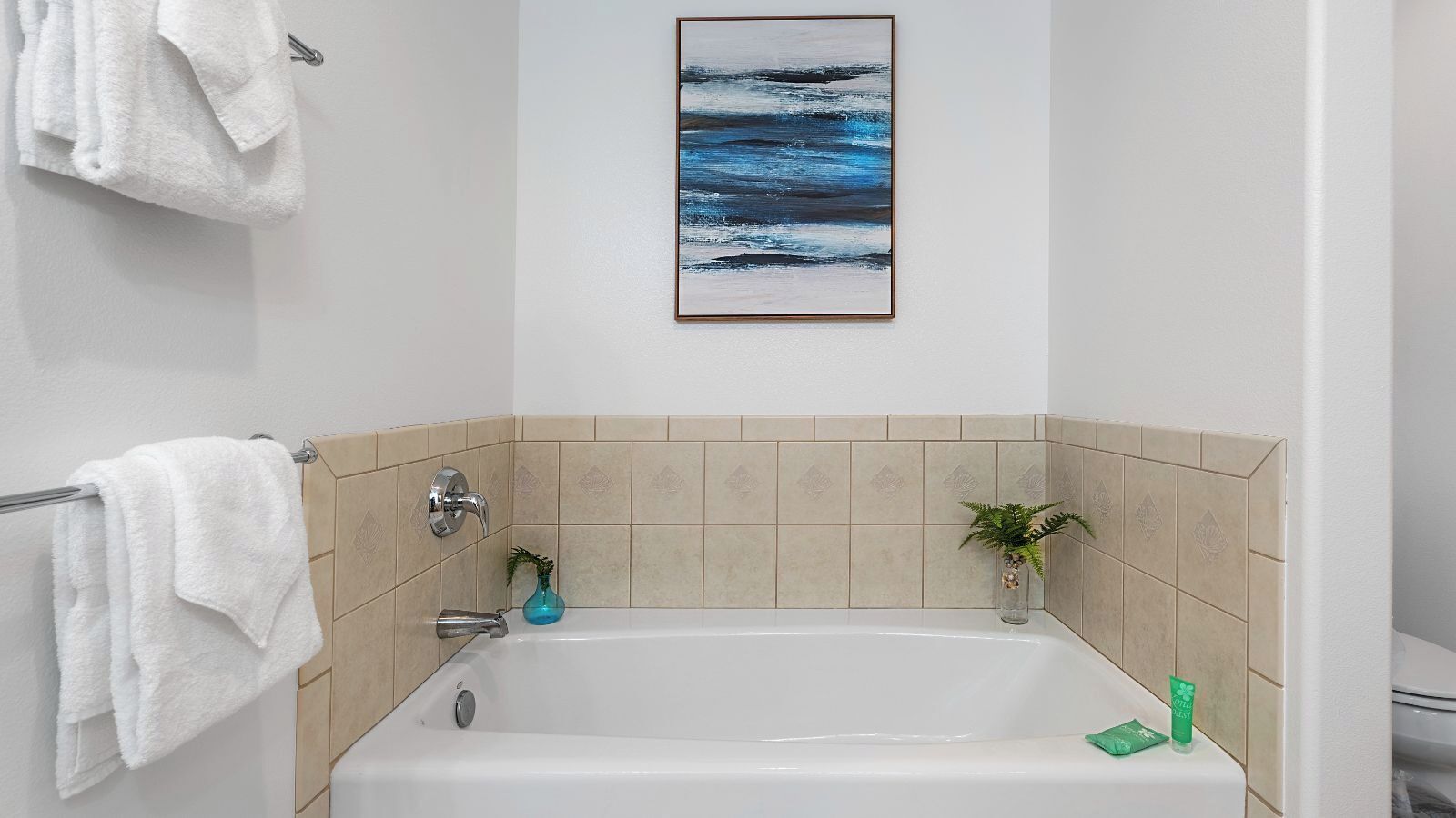 Un baño con bañera, toallas blancas, baldosas color beige, una obra de arte enmarcada y una planta pequeña.
