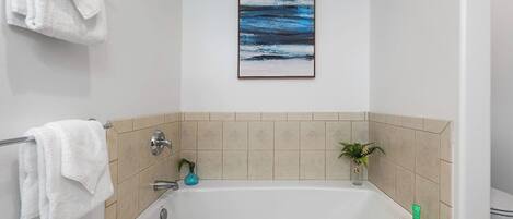 Um banheiro com banheira, toalhas brancas, azulejos bege, uma obra de arte emoldurada e uma pequena planta.