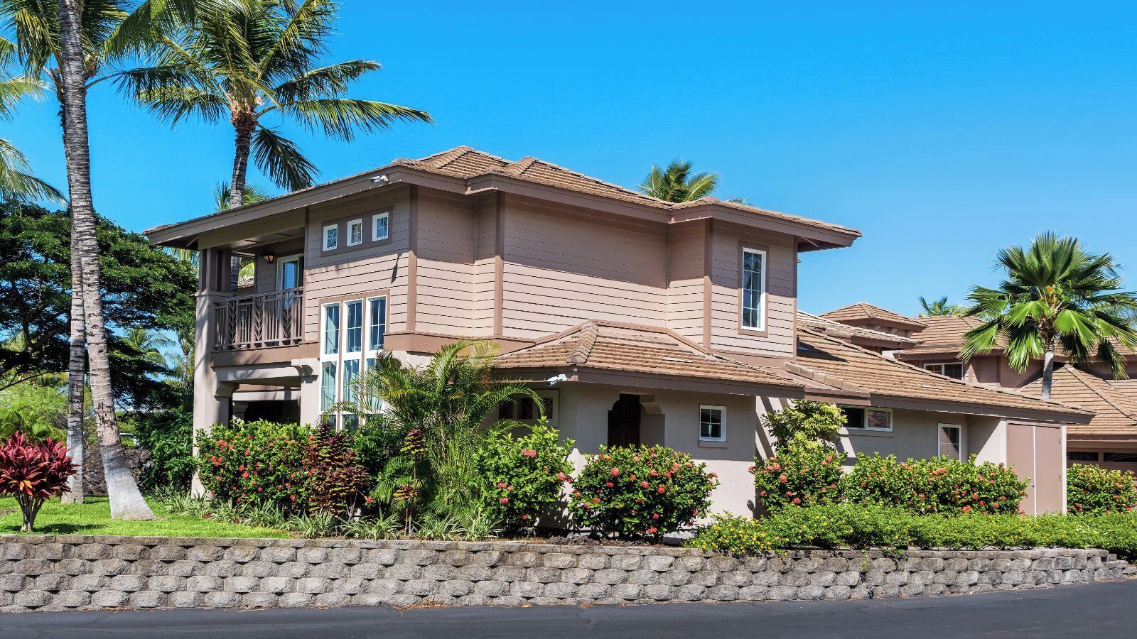 A two-story house with a balcony, surrounded by tropical vegetation and a stone wall.