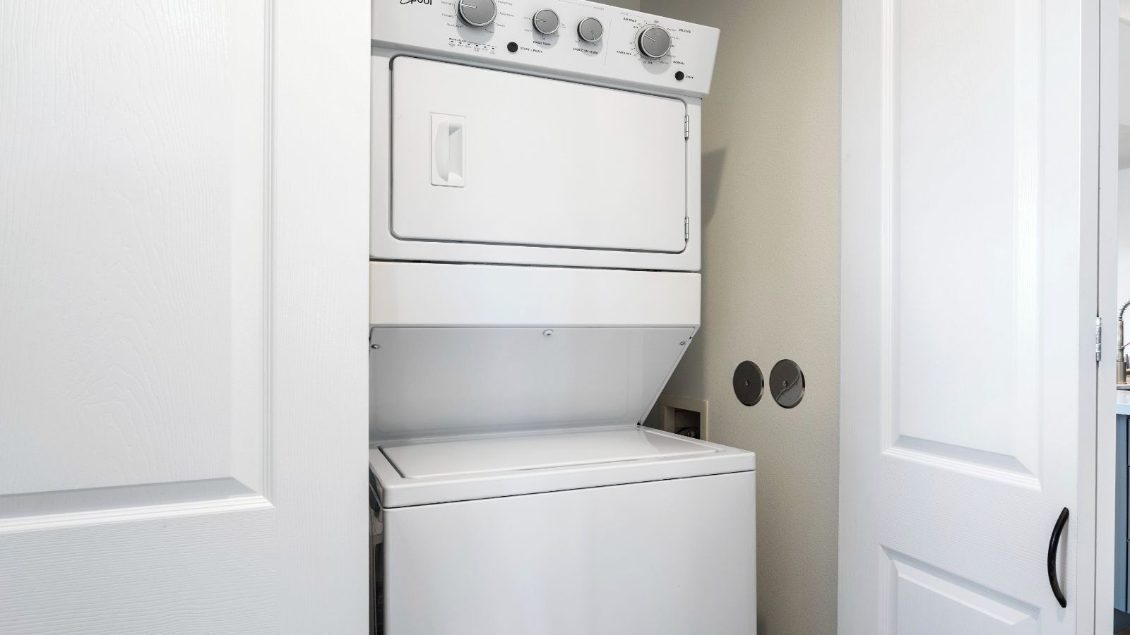 A compact, white laundry room with a front-loading washer and dryer, a control panel with multiple dials, and two wall-mounted circular objects.