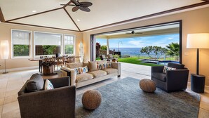 A spacious living area with a large window offering a view of the ocean, a ceiling fan, and a dining area with a table and chairs. - Unbelievable Ocean Front View w Pool - AC - Outdoor Seating!- Vrkcv (Kailua-Kona)