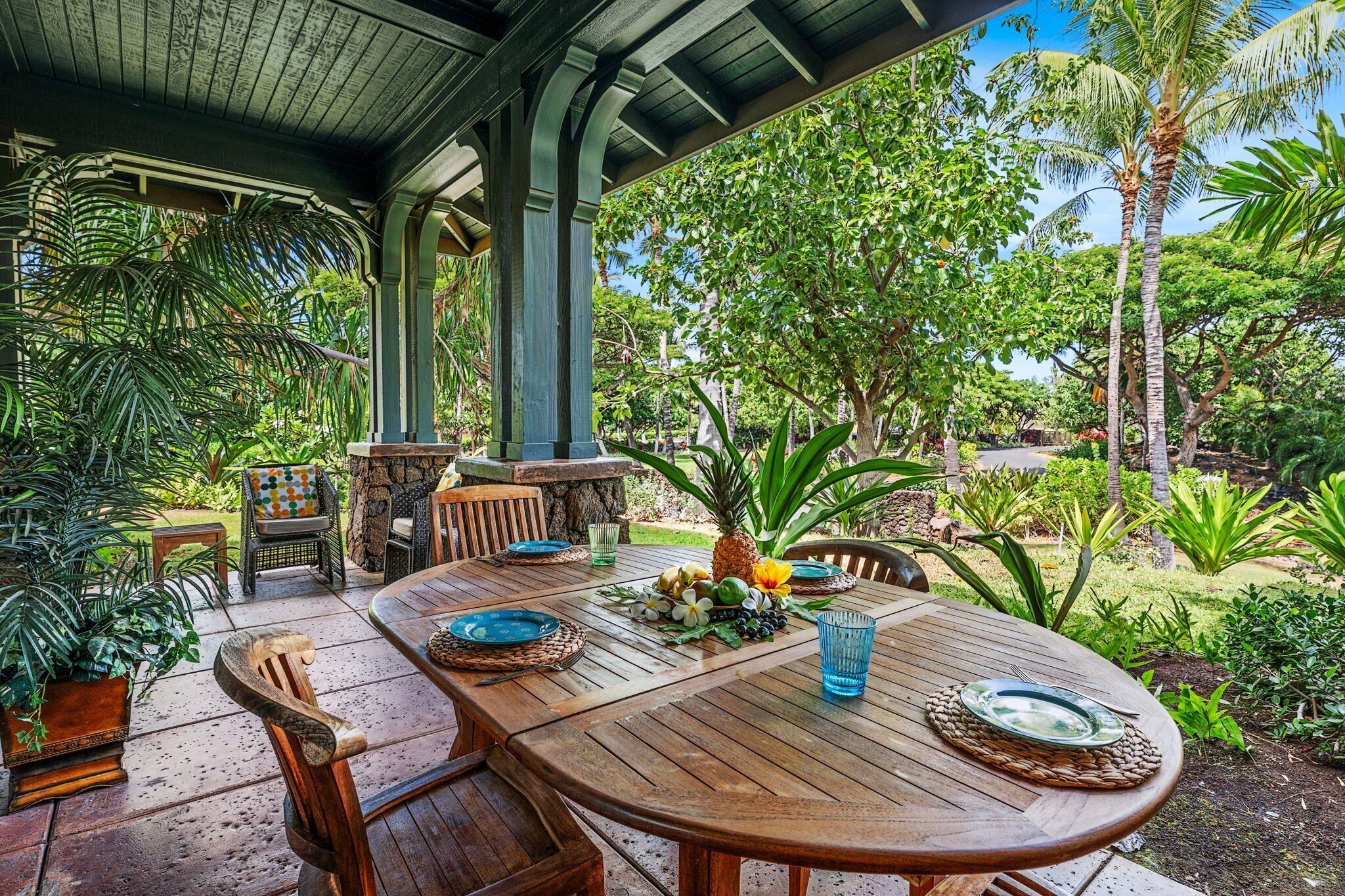 Un espacio exterior para comer con una mesa de madera preparada para dos personas, rodeado de abundante follaje y plantas tropicales.