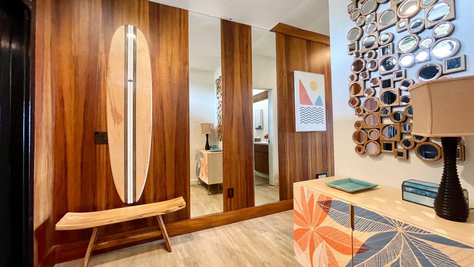 A room with a wooden wardrobe, a surfboard leaning against it, a mirror wall, and a patterned table.