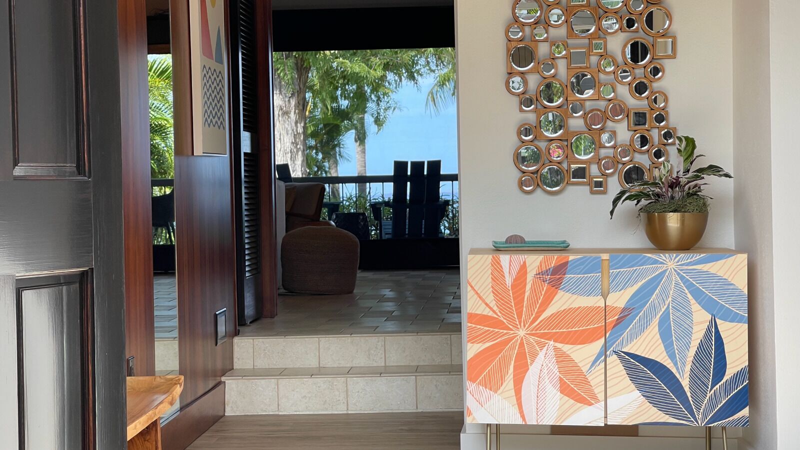 A modern entrance with a wooden door, a tiled floor, a seating area with a chair, a wall decorated with mirrors, and a colorful cabinet with a plant on top.