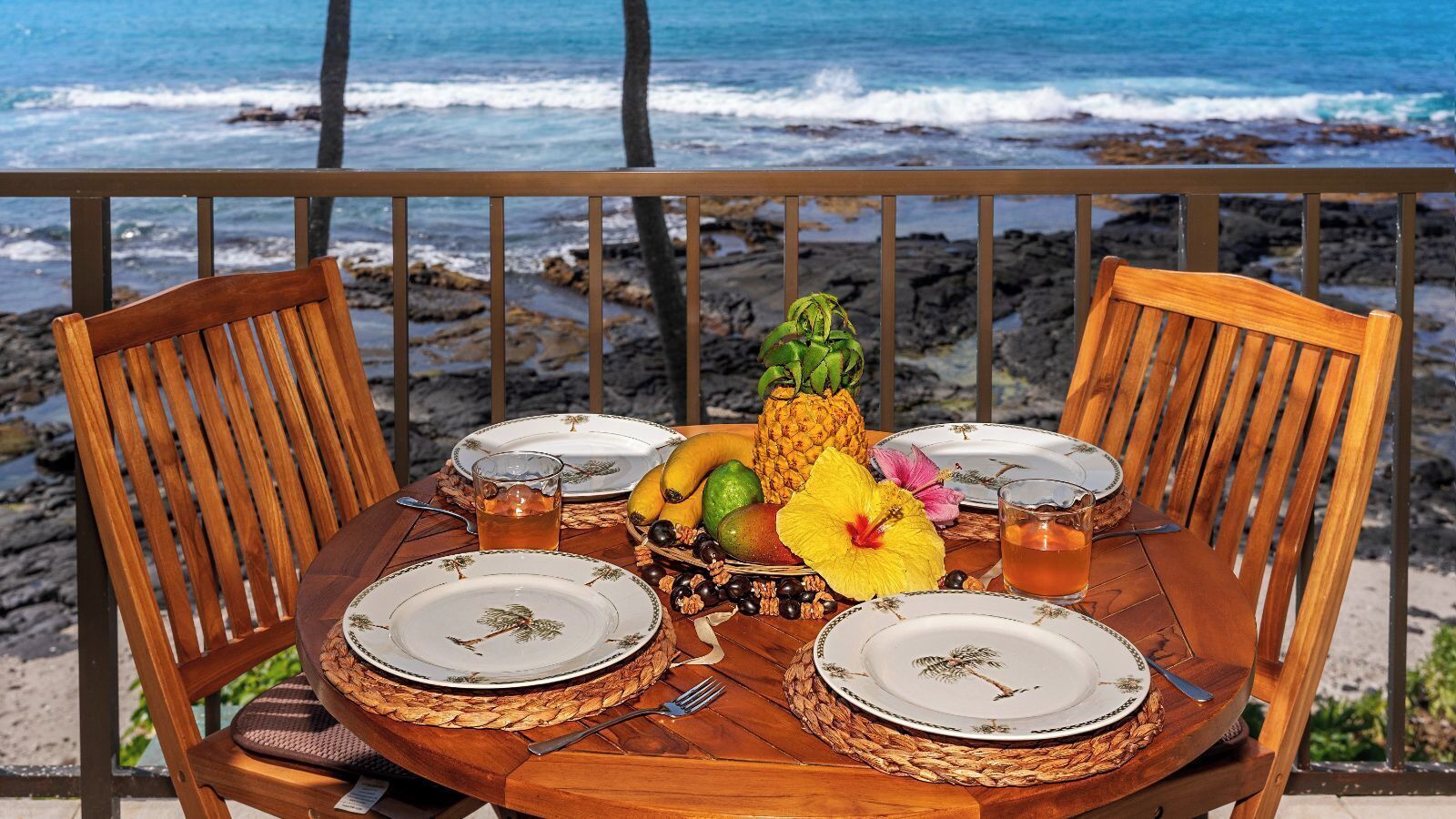 Une table en bois, dressée pour quatre personnes, avec des assiettes, un ananas, des fruits et une fleur d’hibiscus.