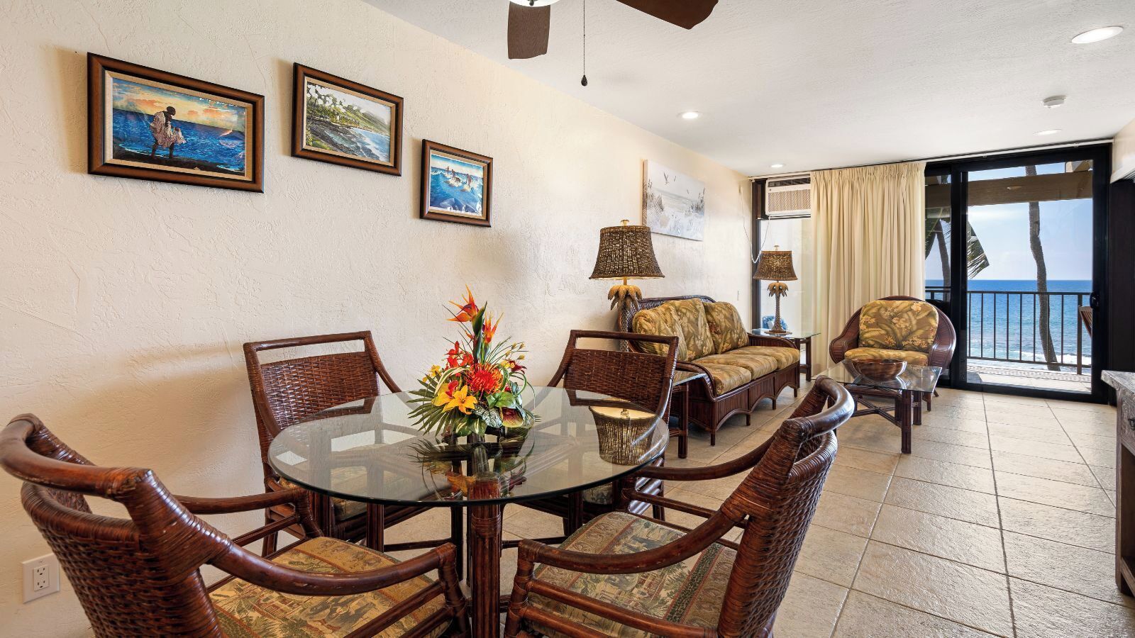 A dining area with a glass table surrounded by wicker chairs, a sofa, and a lamp.