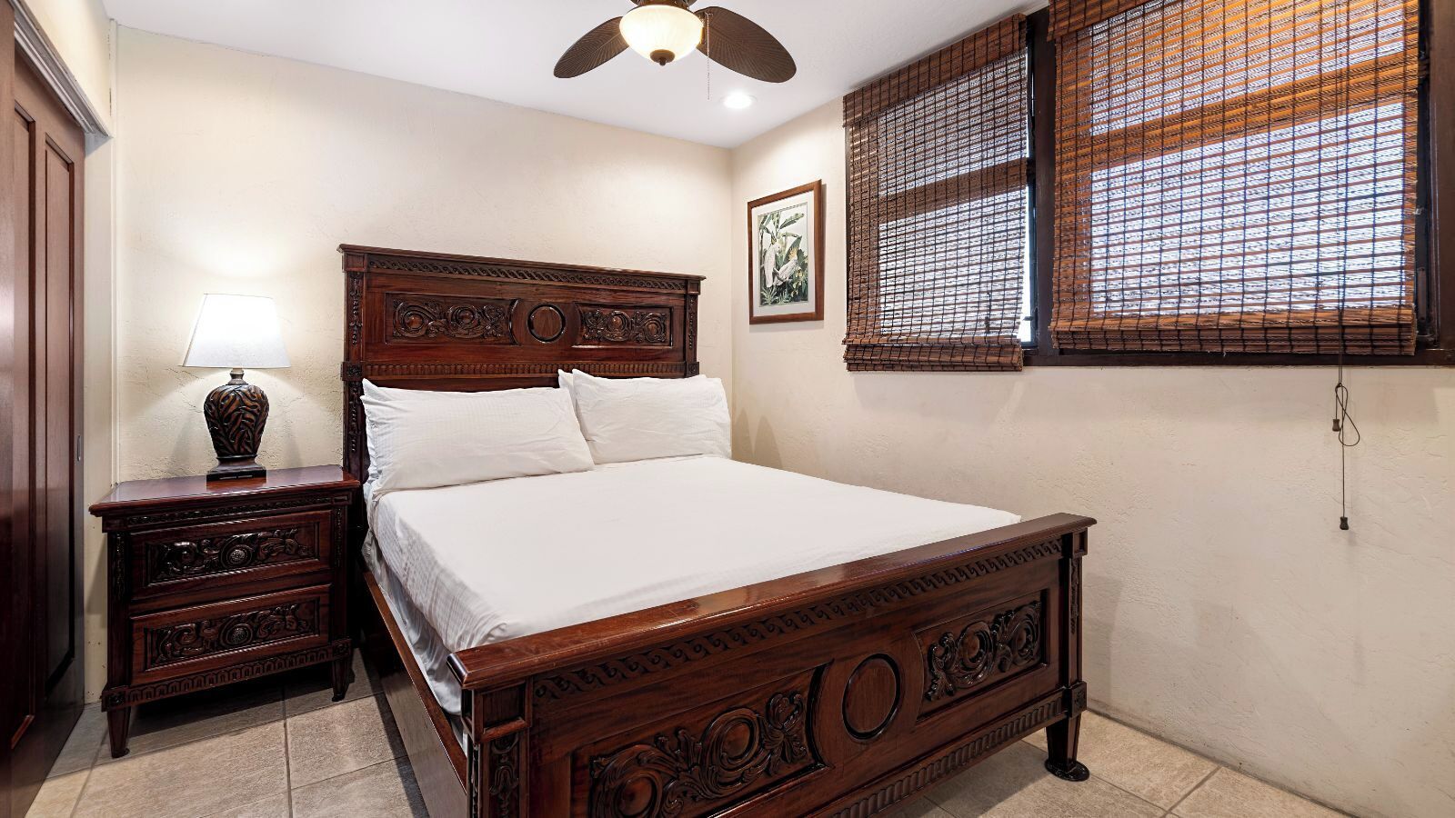 A bedroom with a wooden bed, a nightstand with a lamp, a window with bamboo blinds, and a ceiling fan.