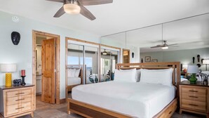A spacious bedroom with a large bed, wooden headboard, and a ceiling fan. There are two nightstands with lamps and a dresser. - Ocean Front Condo - Vrkcv (Kailua-Kona)