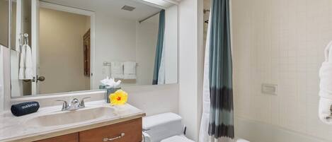 A bathroom with a white sink, mirror, and toilet. There is a towel rack with towels, a makeup bag, and a flower on the sink.