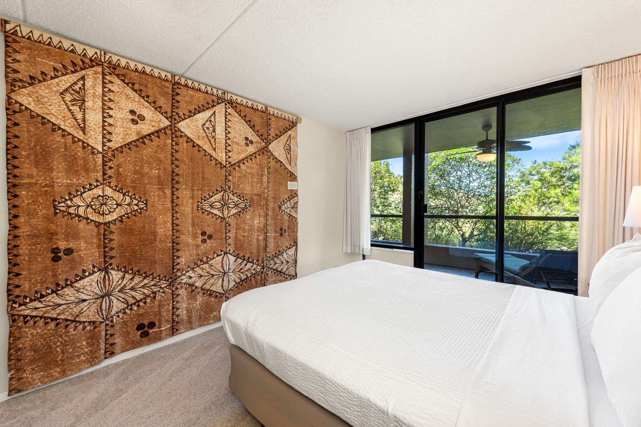 A hotel room with a large bed, a patterned tapestry on the wall, and a view of greenery through the window.