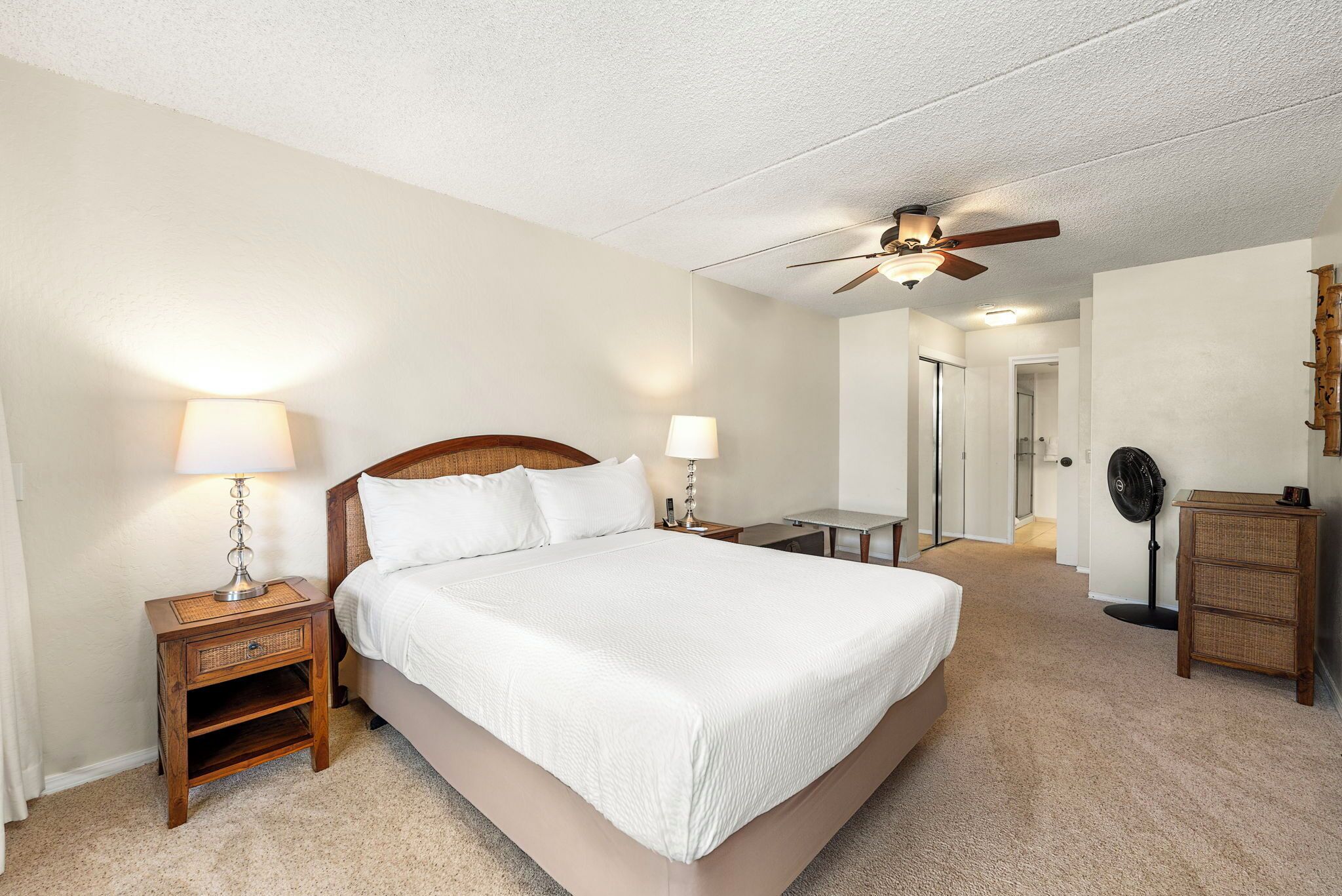 A bedroom with a bed, bedside lamps, a ceiling fan, a desk, and a dresser.