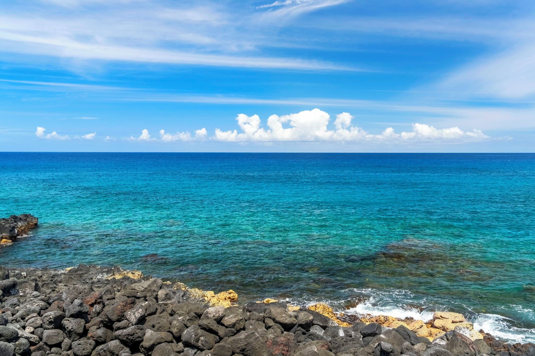 Vista costera con aguas azules cristalinas, un primer plano rocoso y un cielo parcialmente nublado.