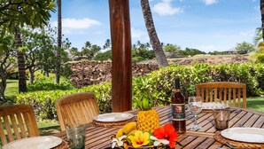 An outdoor dining table with a bottle of Volcano White wine, a pineapple, and tropical fruits, surrounded by wooden chairs and a glass of water, set against a backdrop of palm trees and a clear sky. - Luxurious View Corner Townhome - Great Views - VIP Beach Pass - Vrkcv (Kamuela)