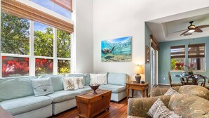 A spacious living room with a light blue sofa, a patterned armchair, a wooden coffee table, and a painting of a mountainous landscape. - Luxurious View Corner Townhome - Great Views - VIP Beach Pass - Vrkcv (Kamuela)