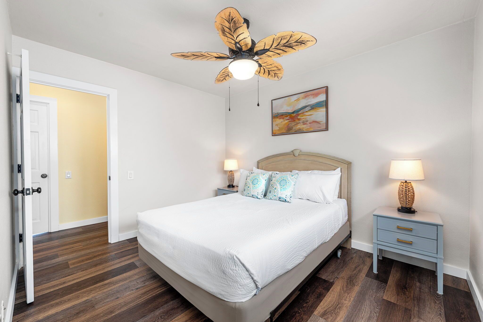 A bedroom with a bed, two bedside tables, a ceiling fan, and a door leading to another room.