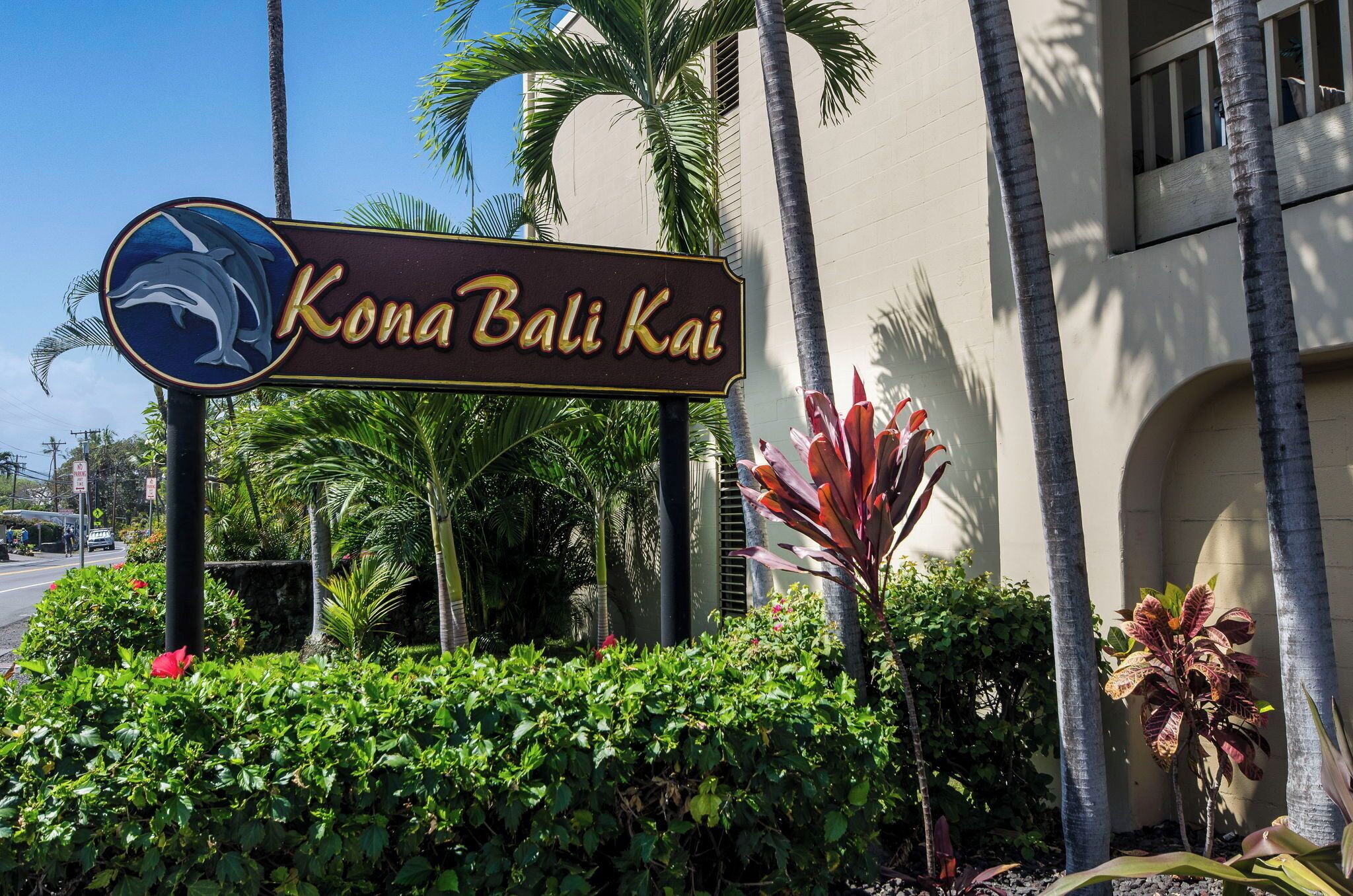 A sign for Kona Bali Kai with a dolphin logo, surrounded by tropical plants and palm trees.