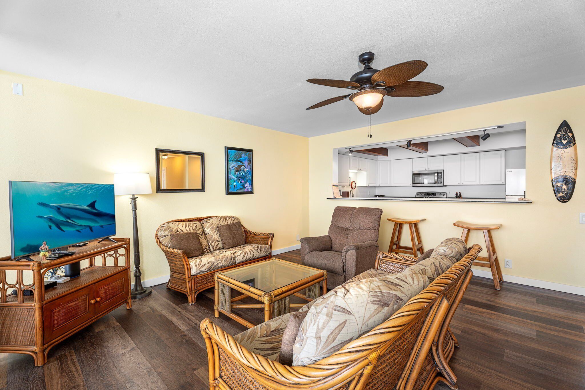 A living room with wicker furniture, a ceiling fan, and a kitchen area visible through an open doorway.