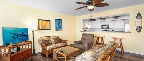 A living room with wicker furniture, a ceiling fan, and a kitchen area visible through an open doorway.