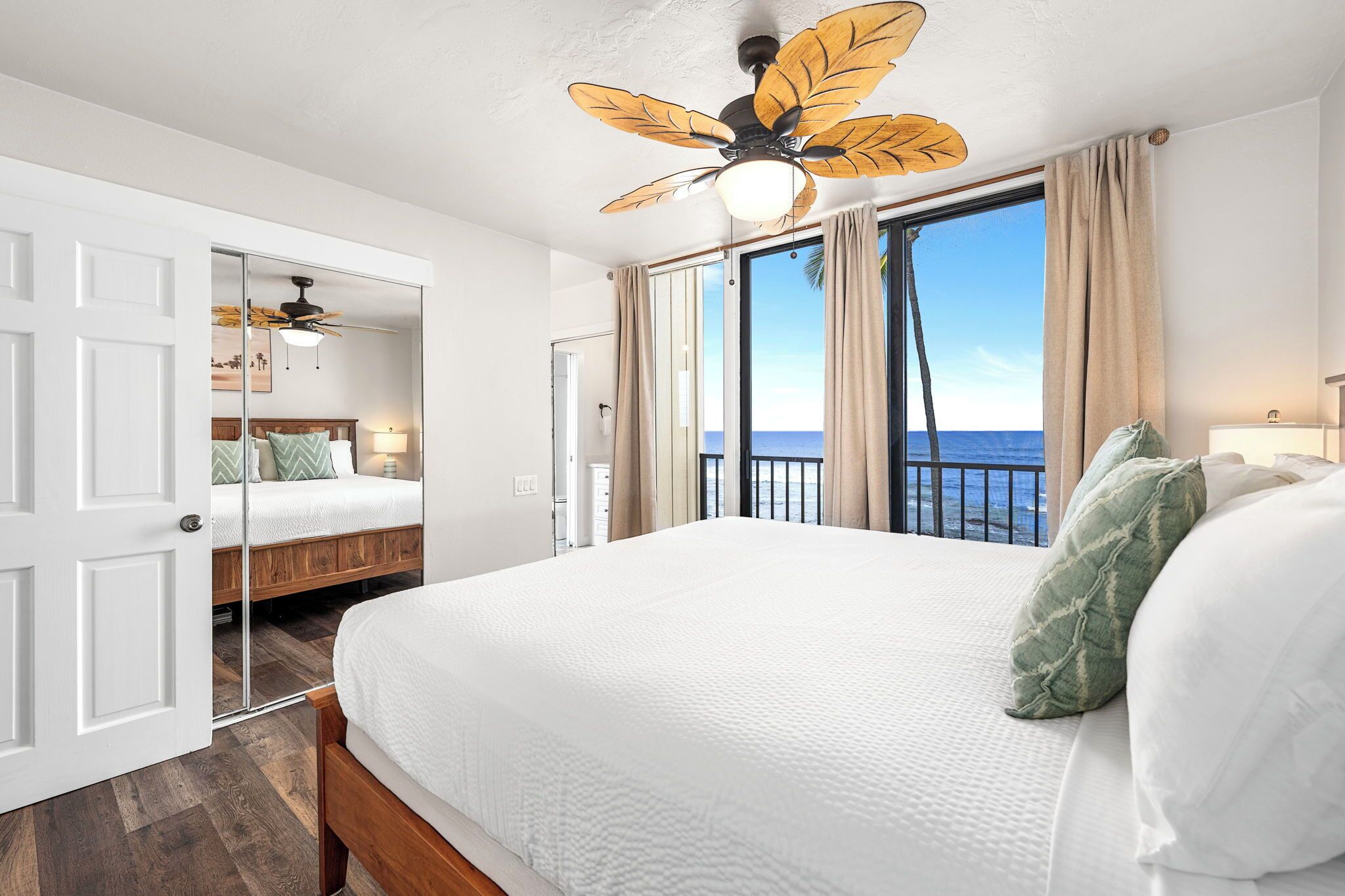 A hotel room with a large bed, a ceiling fan, a view of the ocean, and a balcony.