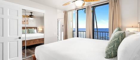 A hotel room with a large bed, a ceiling fan, a view of the ocean, and a balcony.
