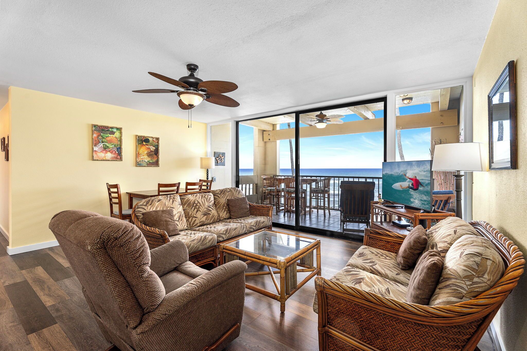 A spacious living area with wicker furniture, a ceiling fan, and a large TV screen displaying a surfer.