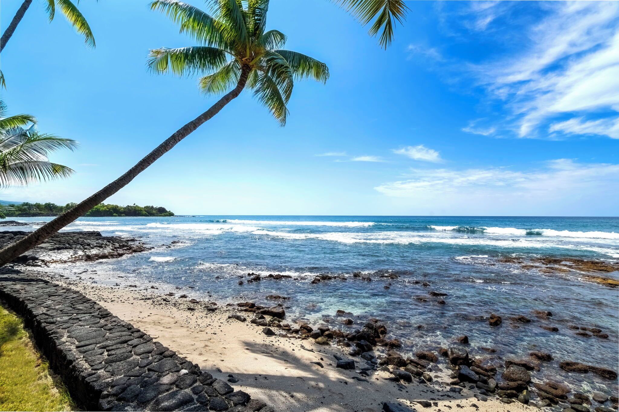 Une plage avec une eau d’un bleu limpide, un mur de pierres et des palmiers.