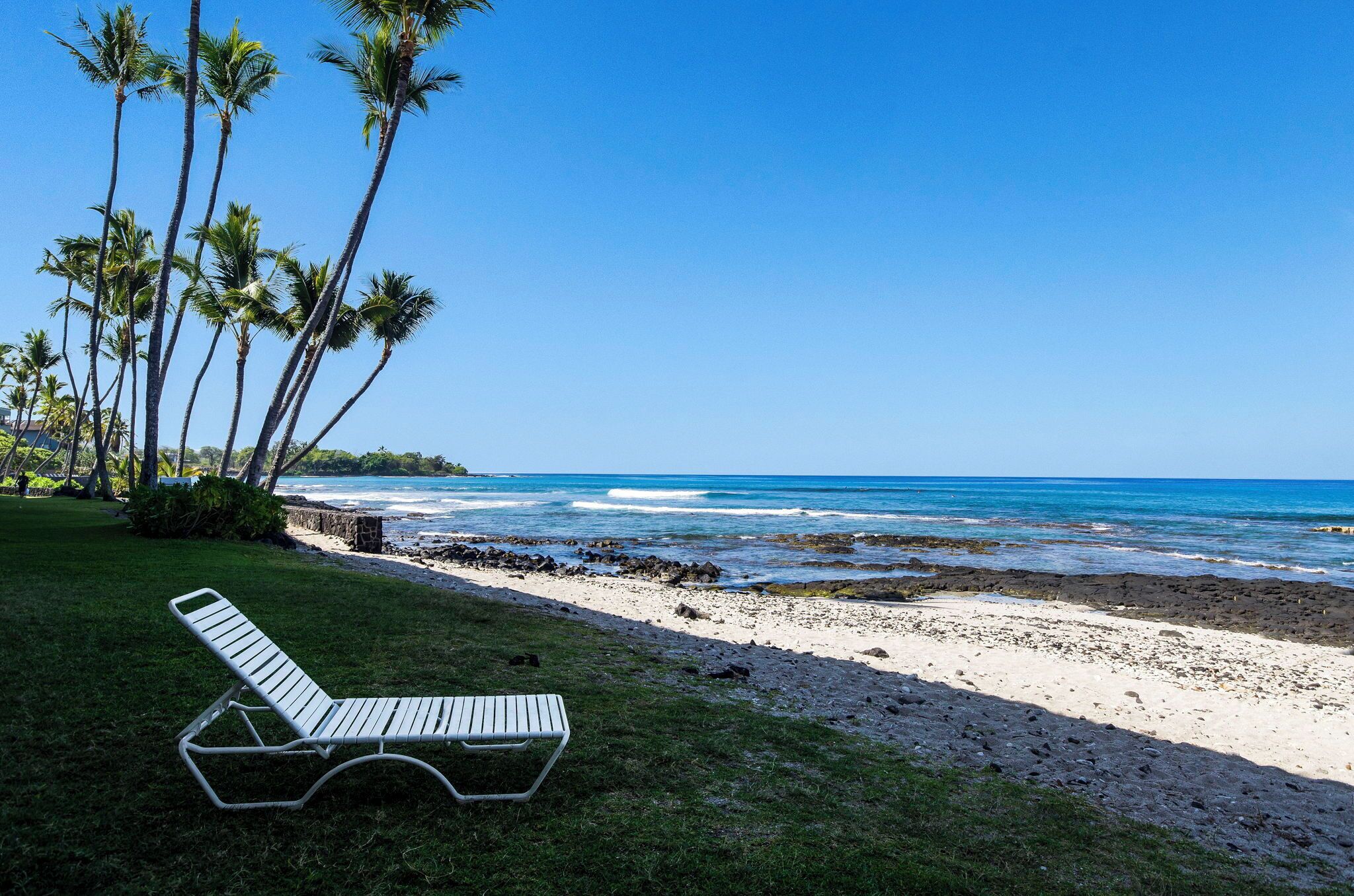 A beach with a lounge chair, palm trees, and ocean waves.