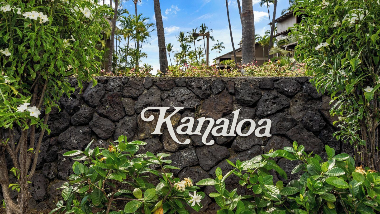 A stone wall with the name "Kanaloa" in cursive lettering, surrounded by lush greenery and palm trees.